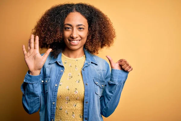 若いですアフリカ系アメリカ人女性とともにアフロ髪身に着けていますカジュアルなデニムシャツ上の黄色の背景ショーと指差し込みで指番号6ながら笑顔自信と幸せ — ストック写真