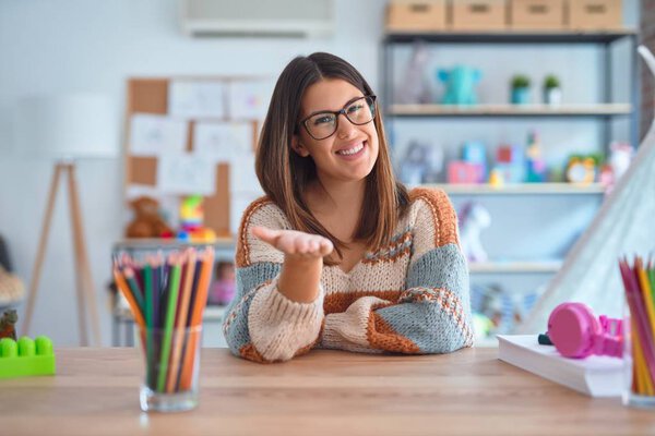 Young beautiful teacher woman wearing sweater and glasses sitting on desk at kindergarten smiling cheerful offering palm hand giving assistance and acceptance.