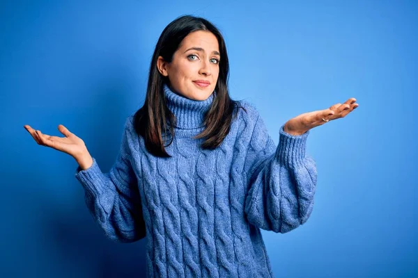 Young brunette woman with blue eyes wearing casual turtleneck sweater clueless and confused expression with arms and hands raised. Doubt concept.