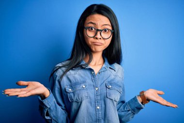 Young beautiful chinese woman wearing casual denim shirt over isolated blue background clueless and confused expression with arms and hands raised. Doubt concept.