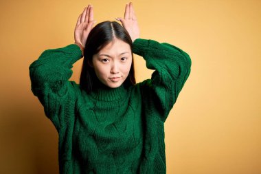 Sarı izole edilmiş arka plan üzerine yeşil kazak giyen genç, güzel Asyalı kadın avuçlarında alaycı ve şüpheci bakışlarla tavşan kulağı hareketi yapıyor. Paskalya tavşanı kavramı.