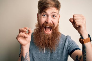 Irish redhead man with beard holding dental invisible aligner for tooth correction screaming proud and celebrating victory and success very excited, cheering emotion