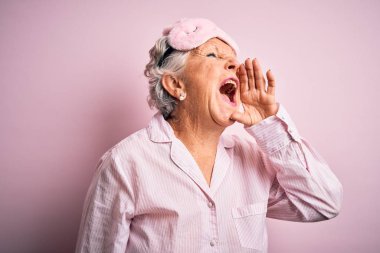 Senior beautiful woman wearing sleep mask and pajama over isolated pink background shouting and screaming loud to side with hand on mouth. Communication concept.