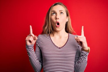 Young beautiful blonde woman wearing casual striped t-shirt over isolated red background amazed and surprised looking up and pointing with fingers and raised arms.