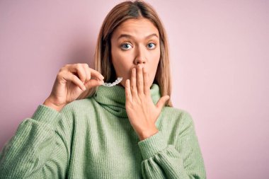 Young beautiful woman holding aligner standing over isolated pink background cover mouth with hand shocked with shame for mistake, expression of fear, scared in silence, secret concept