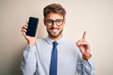 Young businessman holding smartphone standing over isolated white background surprised with an idea or question pointing finger with happy face, number one