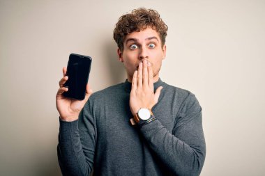 Sarışın, kıvırcık saçlı, akıllı telefonlu, izole edilmiş beyaz arka planda duran genç bir adam. Ağzını kapatıyor. Yanlışlıktan dolayı utancından ağzı açık kalmış, korku ifadesi, sessizlikten korkmak, gizli bir konsept.