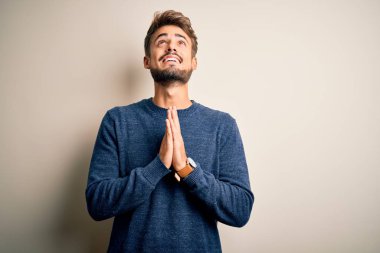 Young handsome man with beard wearing casual sweater standing over white background begging and praying with hands together with hope expression on face very emotional and worried. Begging.