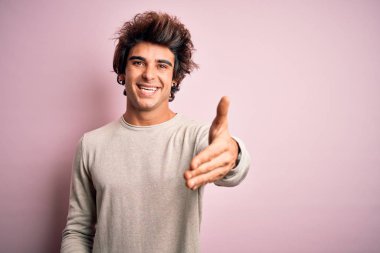 Young handsome man wearing casual t-shirt standing over isolated pink background smiling friendly offering handshake as greeting and welcoming. Successful business.
