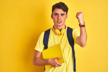 Teenager boy wearing headphones and backpack reading a book over isolated background annoyed and frustrated shouting with anger, crazy and yelling with raised hand, anger concept