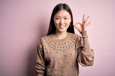Young beautiful asian woman wearing fashion and elegant sweater over pink solated background smiling positive doing ok sign with hand and fingers. Successful expression.