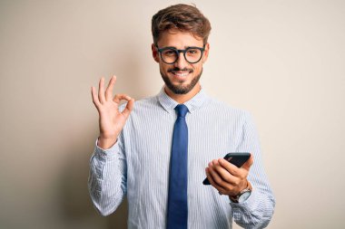 Genç işadamı beyaz arka plan üzerinde akıllı telefon kullanarak konuşuyor. Tamam işareti yapıyor, mükemmel bir sembol.