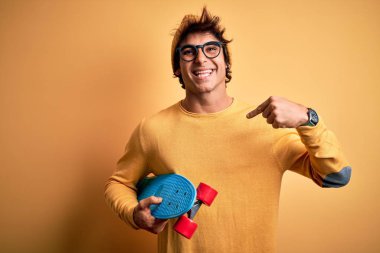 Young handsome student man holding skate wearing glasses over isolated yellow background with surprise face pointing finger to himself