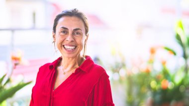 Orta yaşlı güzel bir kadın mutlu ve kendinden emin gülümsüyor terasta yüzünde bir gülümsemeyle duruyor.