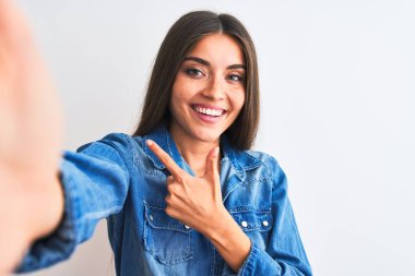 Beautiful woman wearing denim shirt make selfie by camera over isolated white background cheerful with a smile on face pointing with hand and finger up to the side with happy and natural expression