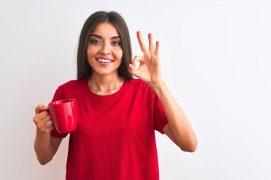 Young beautiful woman drinking red cup of coffee standing over isolated white background doing ok sign with fingers, excellent symbol