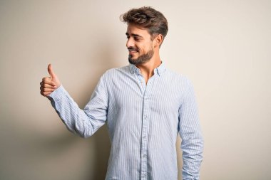 Young handsome man with beard wearing striped shirt standing over white background Looking proud, smiling doing thumbs up gesture to the side