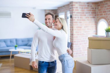 Karton kutular etrafında yeni evde selfie çekmek için akıllı telefon kullanarak ayakta genç güzel çift
