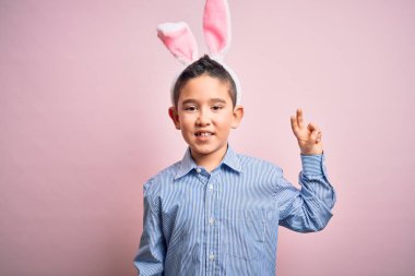 Paskalya tavşanı kulakları takan küçük bir çocuk izole edilmiş pembe arka planda gülümsüyor ve kameraya gülümsüyor zafer işareti yapıyor. İki numara..