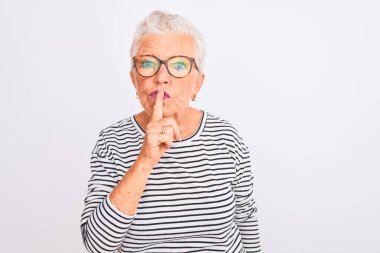 Senior grey-haired woman wearing striped navy t-shirt glasses over isolated white background asking to be quiet with finger on lips. Silence and secret concept.