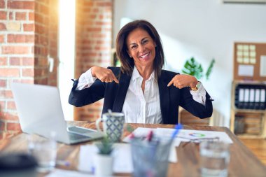 Ofiste dizüstü bilgisayarla çalışan orta yaşlı güzel bir iş kadını yüzünde bir gülümsemeyle gururla ve mutlu bir şekilde kendini işaret ediyor..