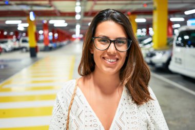 Yeraltı otoparkında araba ve ışıkların etrafında kendine güvenen genç bir kadın.