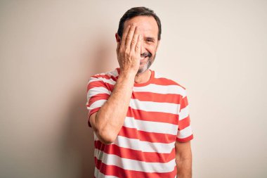 Orta yaşlı, beyaz çizgili tişört giyen bir adam izole edilmiş beyaz arka planda duruyor bir gözünü eliyle kapatıyor, yüzünde kendine güvenen bir gülümseme ve sürpriz bir duygu var..