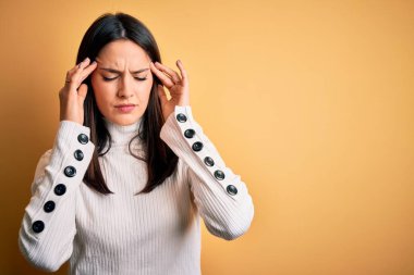 Young brunette woman with blue eyes wearing casual sweater over isolated yellow background suffering from headache desperate and stressed because pain and migraine. Hands on head.