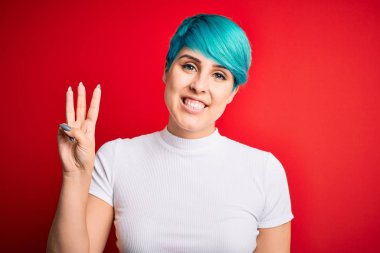 Young beautiful woman with blue fashion hair wearing casual t-shirt over red background showing and pointing up with fingers number three while smiling confident and happy.