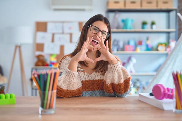 Young beautiful teacher woman wearing sweater and glasses sitting on desk at kindergarten smiling in love doing heart symbol shape with hands. Romantic concept.