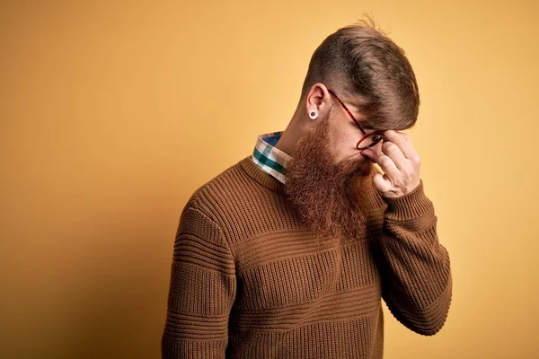 Handsome Irish redhead man with beard wearing glasses and winter sweater over yellow background tired rubbing nose and eyes feeling fatigue and headache. Stress and frustration concept.