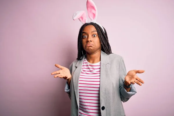 年轻的非洲裔美国女孩 戴着可爱的复活节兔子耳朵 粉红的背景 毫无头绪 双手举着困惑的表情 怀疑的概念 — 图库照片