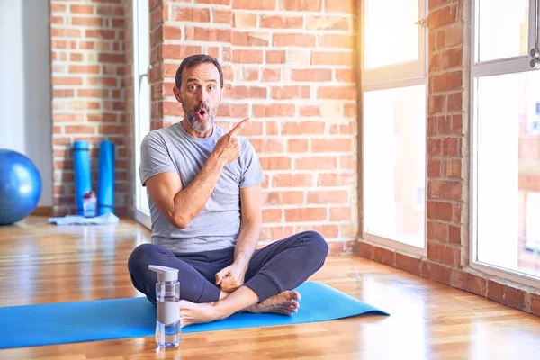 Middle age handsome sportman sitting on mat doing stretching yoga exercise at gym Surprised pointing with finger to the side, open mouth amazed expression.
