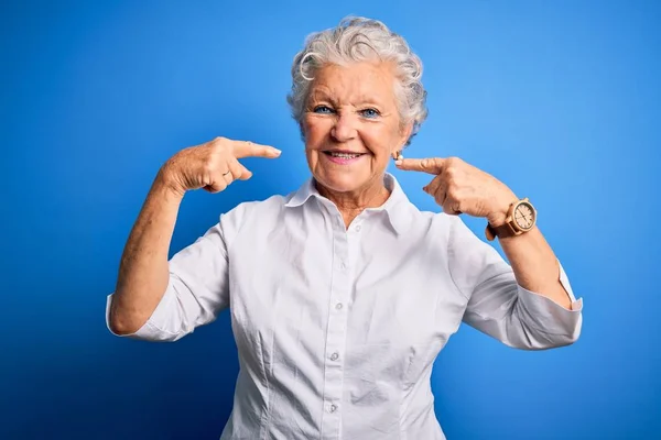 エレガントなシャツを身に着けているシニア美しい女性は 指の歯と口で陽気に示すと指摘笑顔隔離された青の背景の上に立っている 歯科衛生の概念 — ストック写真