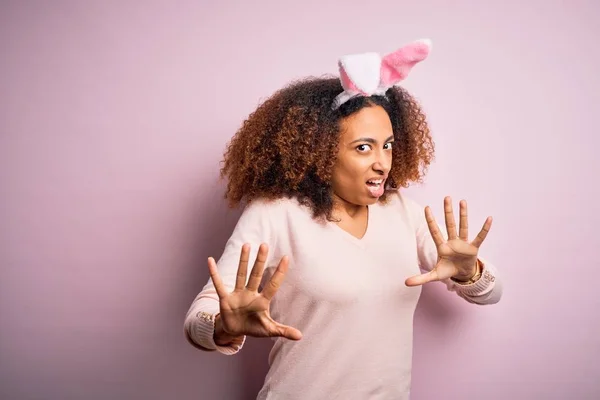 年轻的非洲裔美国女人 黑头发 粉红背景上戴着小兔子耳朵 害怕恐惧的表情 用手停止手势 震惊地大叫 恐慌的概念 — 图库照片
