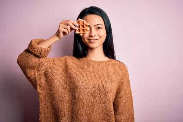 年轻美丽的中国女人拿着华夫饼 站在孤立的粉色背景上 脸上挂着幸福的表情 脸上挂着自信的微笑 露出牙齿 — 图库照片