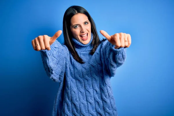 Young brunette woman with blue eyes wearing casual turtleneck sweater approving doing positive gesture with hand, thumbs up smiling and happy for success. Winner gesture.