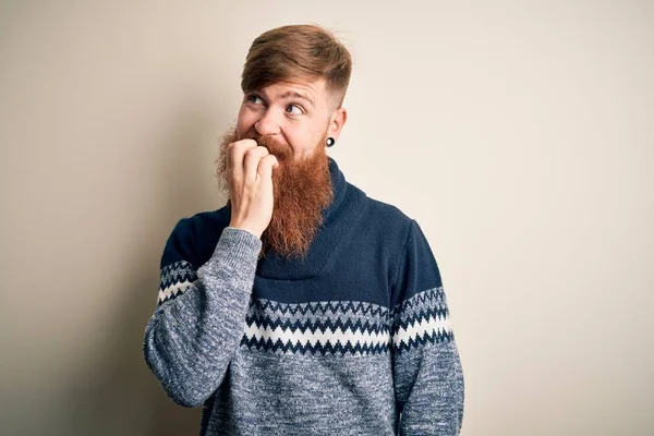 Handsome Irish redhead man with beard wearing winter sweater over isolated background looking stressed and nervous with hands on mouth biting nails. Anxiety problem.