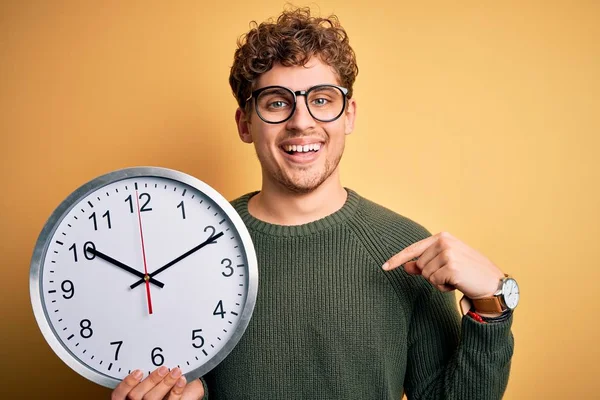 若いですブロンド男とともに巻き毛のある眼鏡をかけ大きな時計上の黄色の背景とともに驚き顔ポインティング指 Gays Tube — ストック写真