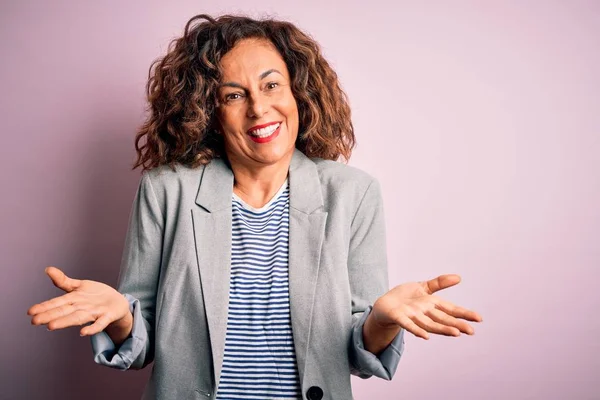 Soyutlanmış pembe arka plan üzerinde zarif bir ceket giyen orta yaşlı güzel bir iş kadını. Kolları ve elleri havada, kafası karışmış bir ifade. Şüphe kavramı.