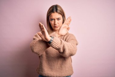 Young beautiful blonde woman wearing winter wool sweater over pink isolated background Rejection expression crossing arms doing negative sign, angry face