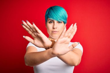 Young beautiful woman with blue fashion hair wearing casual t-shirt over red background Rejection expression crossing arms doing negative sign, angry face