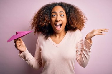Afro saçlı genç Afrikalı bir kadın pembe arka planda kağıt uçak tutuyor çok mutlu ve heyecanlı, kazanan ifade zaferi büyük bir gülümsemeyle kutluyor ve ellerini kaldırıyor.