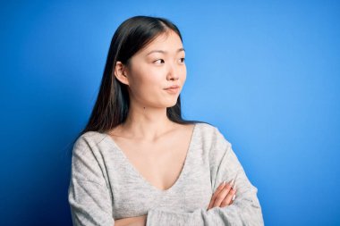 Young beautiful asian woman wearing casual sweater standing over blue isolated background looking to the side with arms crossed convinced and confident