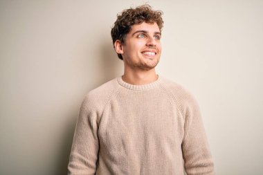 Sarışın, kıvırcık saçlı, beyaz arka planda sıradan bir süveter giyen, yüzünde gülümseme, doğal bir ifade olan genç, yakışıklı bir adam. Kendinden emin gülen.