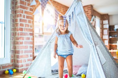 Genç, güzel sarışın kız anaokulunda oyuncaklarla eğleniyor. Evde Kızılderili çadırında oynarken mutlu gülümsüyor.