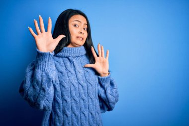 İzole edilmiş mavi arka plan üzerine günlük süveter giyen genç, güzel Çinli kadın korku ifadesinden korkuyor ve dehşete düşüyor. El hareketlerini durdur, şok içinde bağır. Panik konsepti.