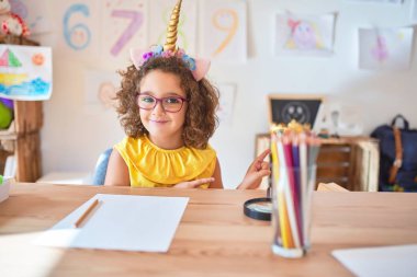 Beautiful toddler wearing glasses and unicorn diadem sitting on desk at kindergarten smiling and looking at the camera pointing with two hands and fingers to the side.