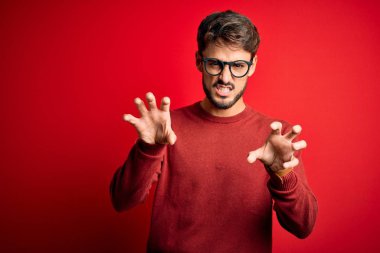 Young handsome man with beard wearing glasses and sweater standing over red background smiling funny doing claw gesture as cat, aggressive and sexy expression