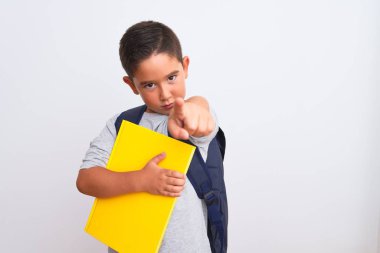 Güzel öğrenci çocuk sırt çantasıyla izole edilmiş beyaz arka plan üzerine kitap tutarak kamerayı işaret ediyor ve sen, el işareti, ön taraftan pozitif ve kendinden emin bir hareket.